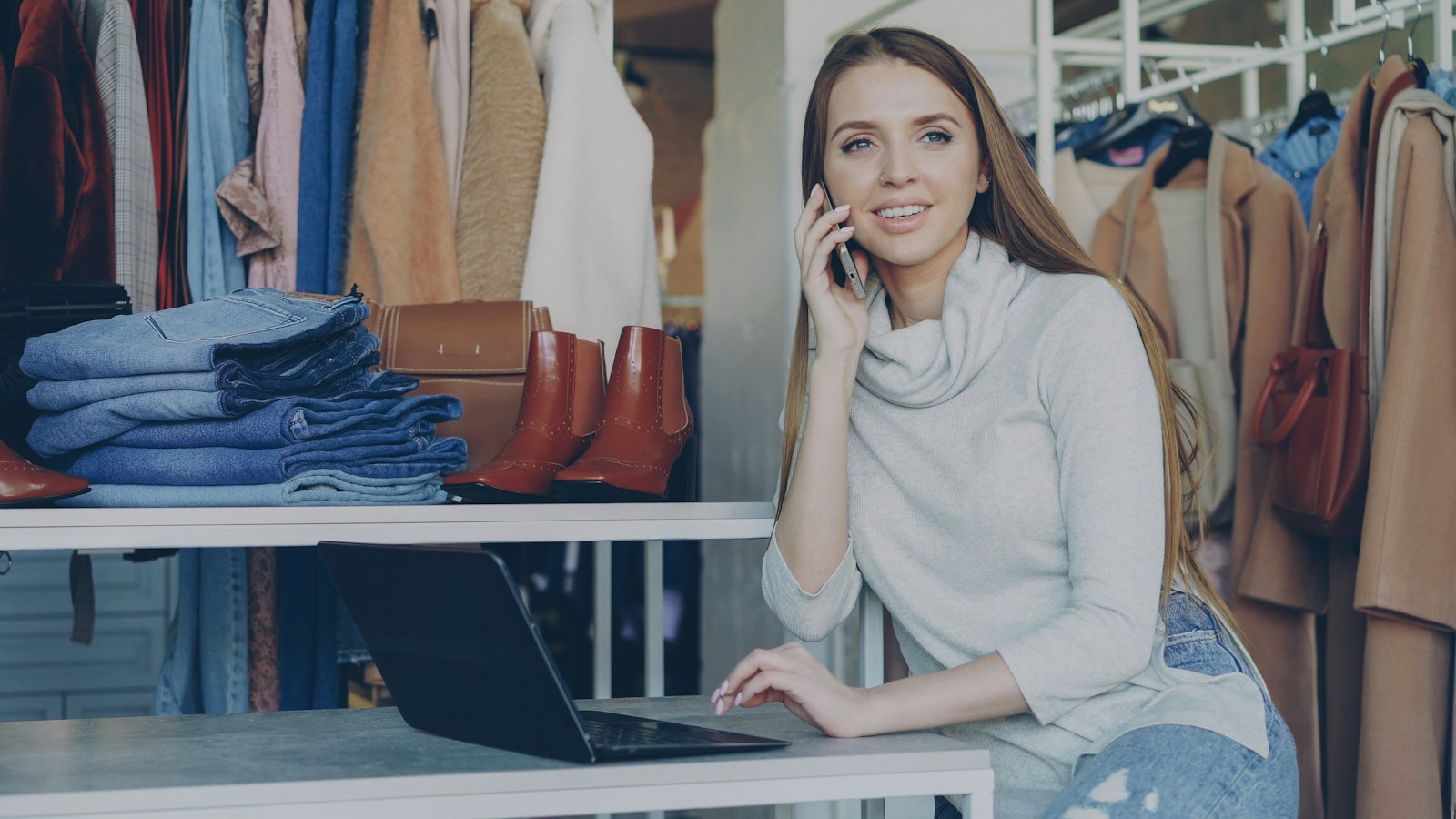 Dona de loja sorrindo enquanto atende ao telefone e mexe no laptop, cercada de roupas