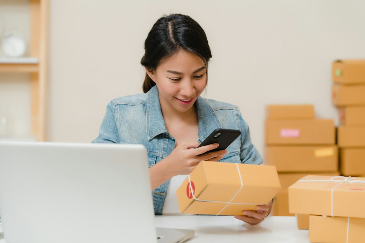 Empreendedora sorrindo enquanto confere um pedido pelo celular, cercada de caixas de mercadoria
